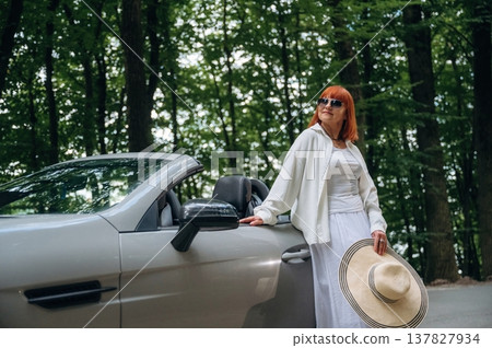 Woman with hat in hand is standing near cabriolet car on the road against trees 137827934