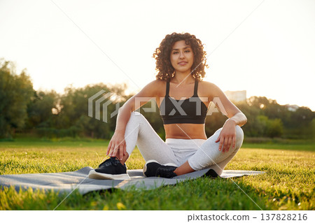 Just resting, sitting on yoga mat and smiling. Young fitness woman is on the field outdoors Just resting, sitting on yoga mat and smiling. Young fitness woman is on the field outdoors 137828216
