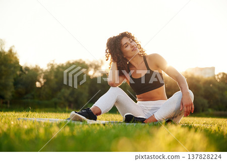 Just resting, sitting on yoga mat and smiling. Young fitness woman is on the field outdoors Just resting, sitting on yoga mat and smiling. Young fitness woman is on the field outdoors 137828224
