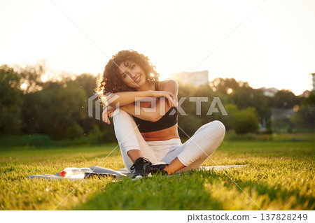 Just resting, sitting on yoga mat and smiling. Young fitness woman is on the field outdoors 137828249