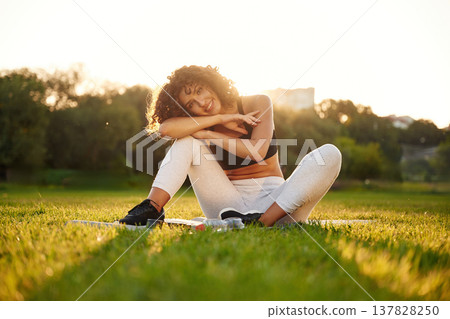 Just resting, sitting on yoga mat and smiling. Young fitness woman is on the field outdoors 137828250