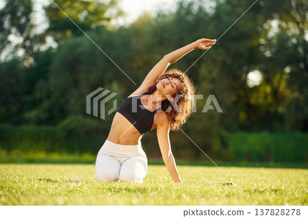 Raising hands up while sitting on the ground. Young fitness woman is on the field outdoors Raising hands up while sitting on the ground. Young fitness woman is on the field outdoors 137828278