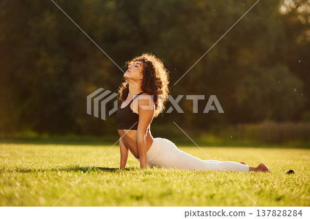 In black top and white pants, doing back stretching. Young fitness woman is on the field outdoors 137828284