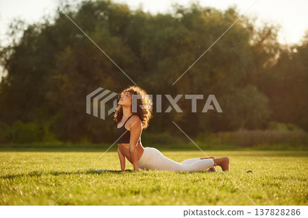 In black top and white pants, doing back stretching. Young fitness woman is on the field outdoors 137828286