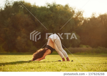 In black top and white pants, doing back stretching. Young fitness woman is on the field outdoors In black top and white pants, doing back stretching. Young fitness woman is on the field outdoors 137828288