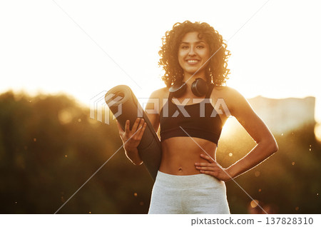 Evening dawn, against sunlight, standing and smiling. Young fitness woman is on the field outdoors Evening dawn, against sunlight, standing and smiling. Young fitness woman is on the field outdoors 137828310