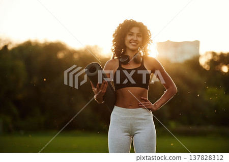 Evening dawn, against sunlight, standing and smiling. Young fitness woman is on the field outdoors Evening dawn, against sunlight, standing and smiling. Young fitness woman is on the field outdoors 137828312
