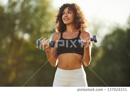 Blue dumbbells are in hands, arms exercise. Young fitness woman is on the field outdoors Blue dumbbells are in hands, arms exercise. Young fitness woman is on the field outdoors 137828331