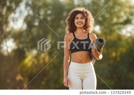 Smiling, holding yoga mat. Young fitness woman is on the field outdoors Smiling, holding yoga mat. Young fitness woman is on the field outdoors 137828349