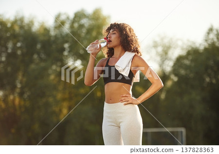 Drinking water from a bottle. Young fitness woman is on the field outdoors Drinking water from a bottle. Young fitness woman is on the field outdoors 137828363