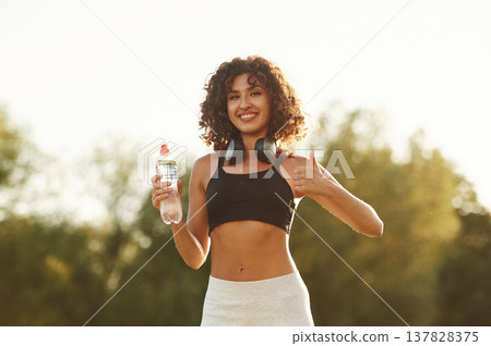 Headphones on neck, bottle of water in hand. Young fitness woman is on the field outdoors Headphones on neck, bottle of water in hand. Young fitness woman is on the field outdoors 137828375