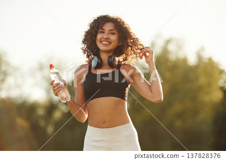 Headphones on neck, bottle of water in hand. Young fitness woman is on the field outdoors Headphones on neck, bottle of water in hand. Young fitness woman is on the field outdoors 137828376