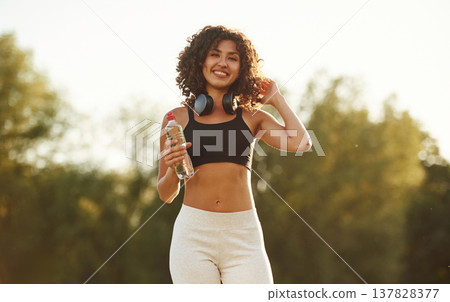 Headphones on neck, bottle of water in hand. Young fitness woman is on the field outdoors Headphones on neck, bottle of water in hand. Young fitness woman is on the field outdoors 137828377