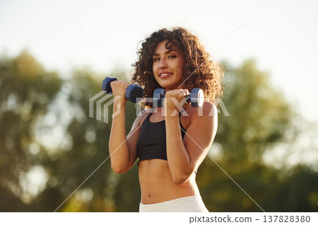 Lifting the dumbbells. Young fitness woman is on the field outdoors Lifting the dumbbells. Young fitness woman is on the field outdoors 137828380