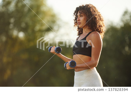 Blue dumbbells are in hands, arms exercise. Young fitness woman is on the field outdoors Blue dumbbells are in hands, arms exercise. Young fitness woman is on the field outdoors 137828394