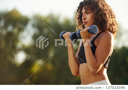 Blue dumbbells are in hands, arms exercise. Young fitness woman is on the field outdoors Blue dumbbells are in hands, arms exercise. Young fitness woman is on the field outdoors 137828396