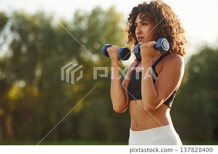 Weight lifting, dumbbells in hands. Young fitness woman is on the field outdoors 137828400