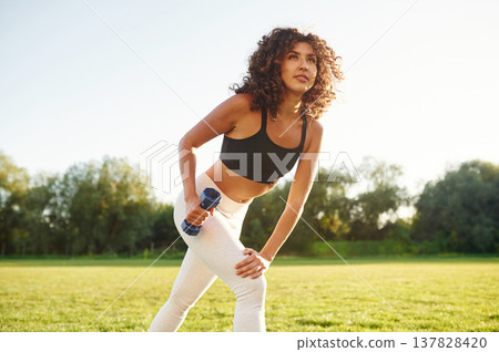 Doing lifting exercises by using dumbbell. Young fitness woman is on the field outdoors Doing lifting exercises by using dumbbell. Young fitness woman is on the field outdoors 137828420