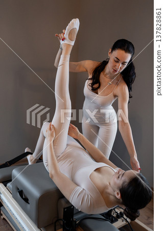 Professional pilates trainer helping woman hold legs up for advanced flexibility on mat in sunlit room. Leg up, pilates trainer, advanced, mat, flexibility. Professional pilates trainer helping woman hold legs up for advanced flexibility on mat in sunlit room. Leg up, pilates trainer, advanced, mat, flexibility. 137828861