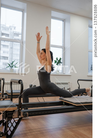 Woman doing pilates challenge raising arms upright on reformer in minimal white office setting. Pilates challenge, raising arms, reformer, white setting, upright arms. 137828886