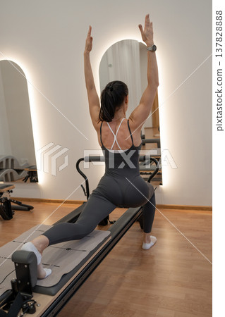 Woman in black lifting arms high on pilates reformer, side view in modern studio with light wall. Lifting arms, pilates reformer, modern studio, black outfit. 137828888