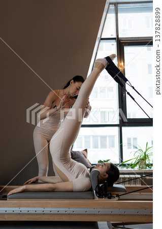 Coach helps woman with stretching legs on pilates machine in modern sunlit fitness room. Stretching legs, pilates, machine, coach, sunlit. Coach helps woman with stretching legs on pilates machine in modern sunlit fitness room. Stretching legs, pilates, machine, coach, sunlit. 137828899
