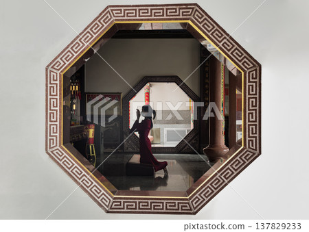 Through the etched geometry of an octagonal portal, the world falls away into a hushed sanctuary of crimson and shadow. There, a woman kneels in a profoundly intimate act of devotion, her vibrant 137829233