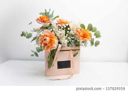 Orange rose bouquet with eucalyptus branches and delicate white flowers arranged in a pastel gift box. Floristry, celebration, romance, decorative composition, and elegant botanical design for festive 137829359