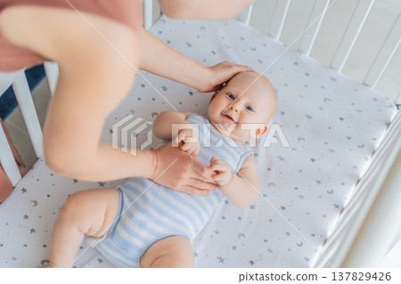 Mother sitting beside the crib gently stroking her baby lying in a cozy bed at home. Warm intimate moment filled with love, trust and calm. Motherhood, infant comfort and emotional bonding concept Mother sitting beside the crib gently stroking her baby lying in a cozy bed at home. Warm intimate moment filled with love, trust and calm. Motherhood, infant comfort and emotional bonding concept 137829426