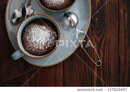 Two chocolate mug cake under sugar icing on dark wooden table. Easy homemade dessert. Mugcake from microwave. Top view Two chocolate mug cake under sugar icing on dark wooden table. Easy homemade dessert. Mugcake from microwave. Top view 137829497