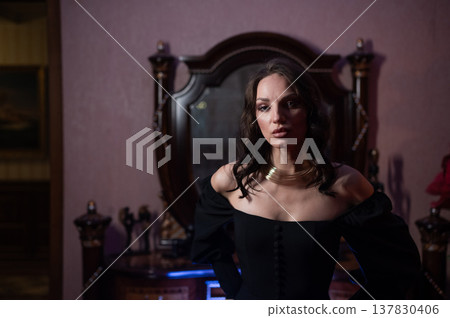 A beautiful brunette in a black evening dress poses in front of a retro-style mirror. A beautiful brunette in a black evening dress poses in front of a retro-style mirror. 137830406