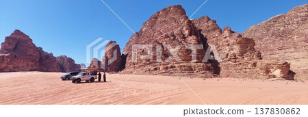 Panoramic view of a desert landscape with towering sandstone cliffs, rippled sand dunes and Off-road vehicles 137830862