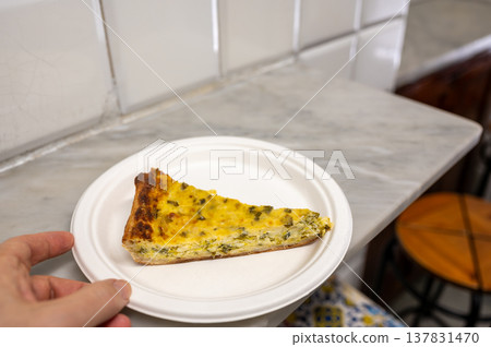 Hand holding sliced traditional ligurian savory pie on a compostable plate, showcasing regional italian street food gastronomy against a tiled wall backdrop Hand holding sliced traditional ligurian savory pie on a compostable plate, showcasing regional italian street food gastronomy against a tiled wall backdrop 137831470