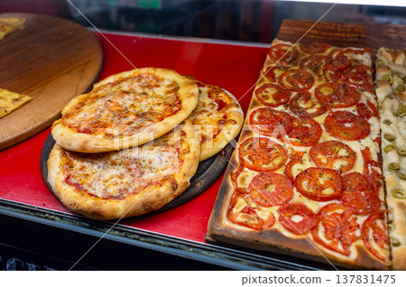 Displaying traditional ligurian baked goods including round pizzas with melted cheese and rectangular focaccia topped with fresh tomatoes and green olives ready for sale in a bakery Displaying traditional ligurian baked goods including round pizzas with melted cheese and rectangular focaccia topped with fresh tomatoes and green olives ready for sale in a bakery 137831475