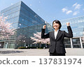 A Japanese woman in a business suit stands in front of a company building amidst falling cherry blossoms, steeling herself for the occasion. Image of a new employee (AI generated). 137831974