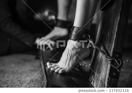 A man chains a woman's legs to a BDSM cross. A man chains a woman's legs to a BDSM cross. 137832116