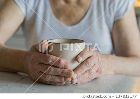 Senior woman in white shirt holding a warm ceramic mug with both wrinkled hands on a white wooden table, peaceful routine Senior woman in white shirt holding a warm ceramic mug with both wrinkled hands on a white wooden table, peaceful routine 137832177