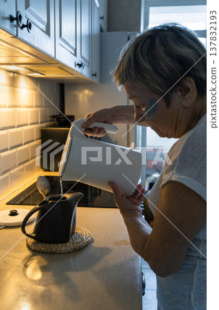 An elderly woman with dark circles under her eyes pours hot water from an electric kettle into teapot in her kitchen, enjoying her morning routine. Real people, life of an elderly person 137832193