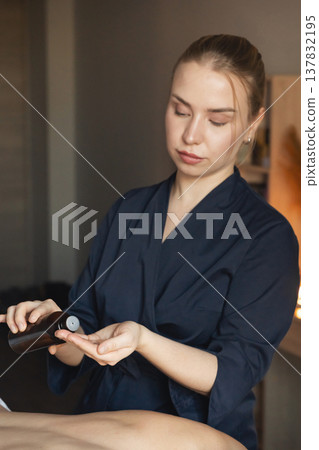 Professional masseuse pouring essential oil into hand, preparing for a calming body massage and wellness therapy session Professional masseuse pouring essential oil into hand, preparing for a calming body massage and wellness therapy session 137832195