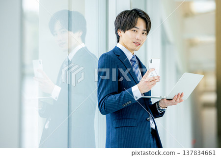 一位身穿西裝的年輕商務人士站在辦公室裡，面前擺放著一台筆記型電腦。 137834661