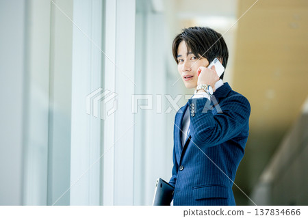 A smiling young man talking on his smartphone in the office lobby. A smiling young man talking on his smartphone in the office lobby. 137834666