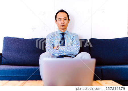 A Japanese man is lost in thought, sitting on his PC on the sofa. 137834879