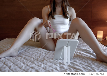 A young woman performs self-care on her VIO (bikini area) in the groin area, shaving and removing hair with a T-shaped razor while looking in a mirror. A young woman performs self-care on her VIO (bikini area) in the groin area, shaving and removing hair with a T-shaped razor while looking in a mirror. 137836761