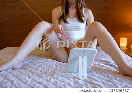 A young woman performs self-care on her VIO (vaginal, inner thigh) area, shaving with a razor while looking in a mirror. 137836787
