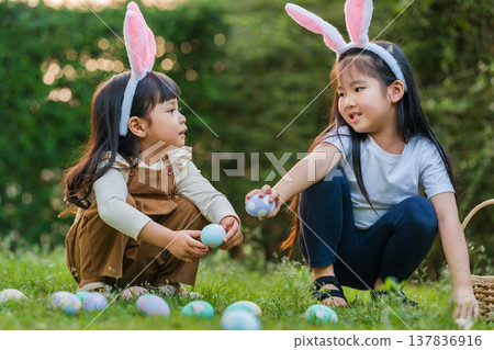 two child girl wearing bunny ears playing together and hunting for Easter eggs into basket on green grass in garden two child girl wearing bunny ears playing together and hunting for Easter eggs into basket on green grass in garden 137836916