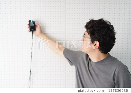 A bespectacled man uses a light meter to measure the intensity of light in a room lined with sound-absorbing boards. A bespectacled man uses a light meter to measure the intensity of light in a room lined with sound-absorbing boards. 137836986