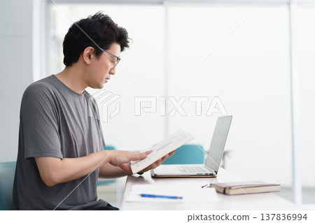 一位三十多歲的日本男子，戴著眼鏡，正在教室裡認真地閱讀文件，他的電腦和書寫工具都觸手可及。 137836994
