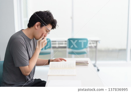 A bespectacled man in his 30s is intently reading textbooks and reference books in a classroom. A bespectacled man in his 30s is intently reading textbooks and reference books in a classroom. 137836995