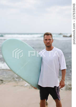 Man Wearing White Shirt Holds Surfboard. Frontfacing Male Model With Pastel Surfboard In Bright Setting Man Wearing White Shirt Holds Surfboard. Frontfacing Male Model With Pastel Surfboard In Bright Setting 137838048