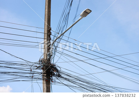 Messy tangled electrical cables and telecommunication network wires hanging chaotically on a concrete utility pole with an LED street light against a clear blue sky. Urban infrastructure hazard. 137838232